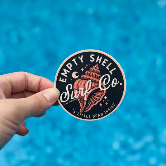 Hand holding a 'Empty Shell Surf Co.' sticker against a blue water background