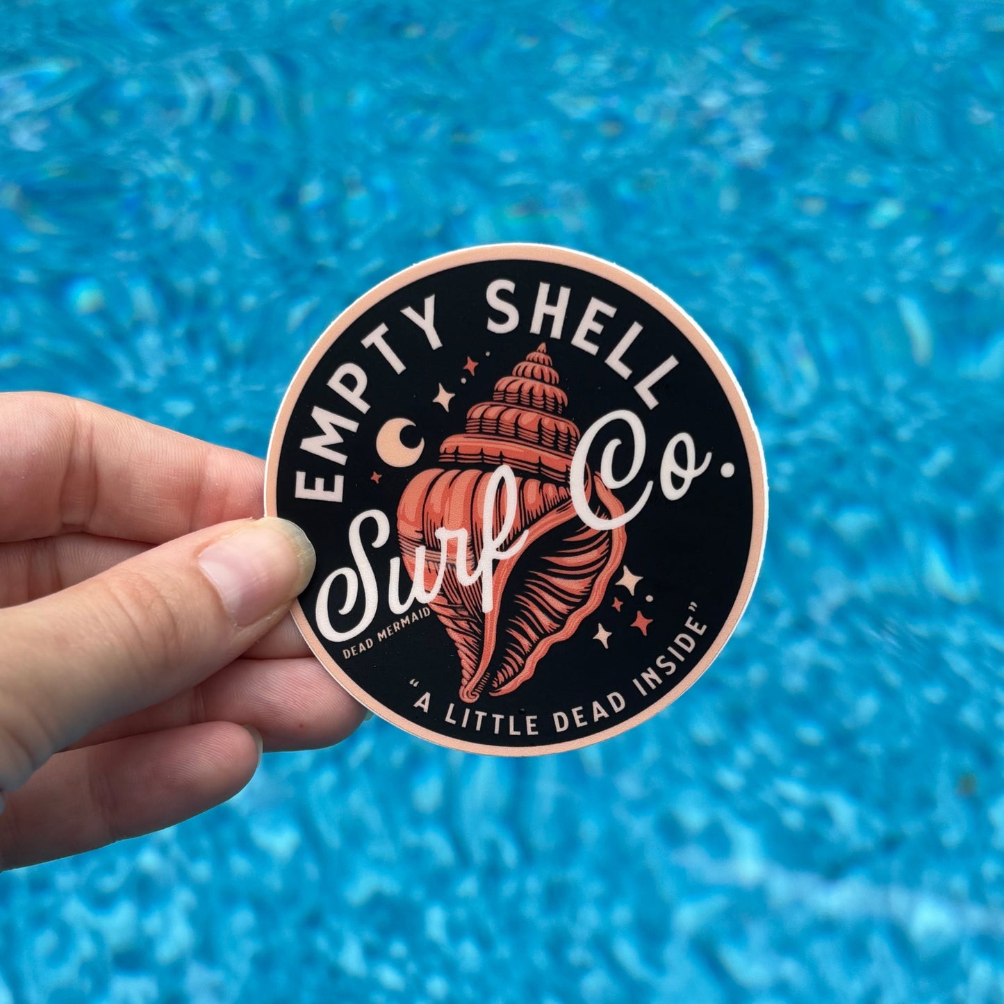 Hand holding a circular sticker with a shell design against a blue water background