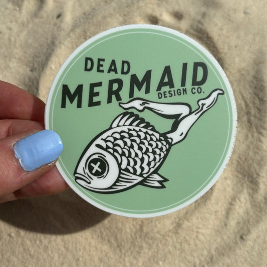 Green sticker with a mermaid design held by a hand on a sandy background