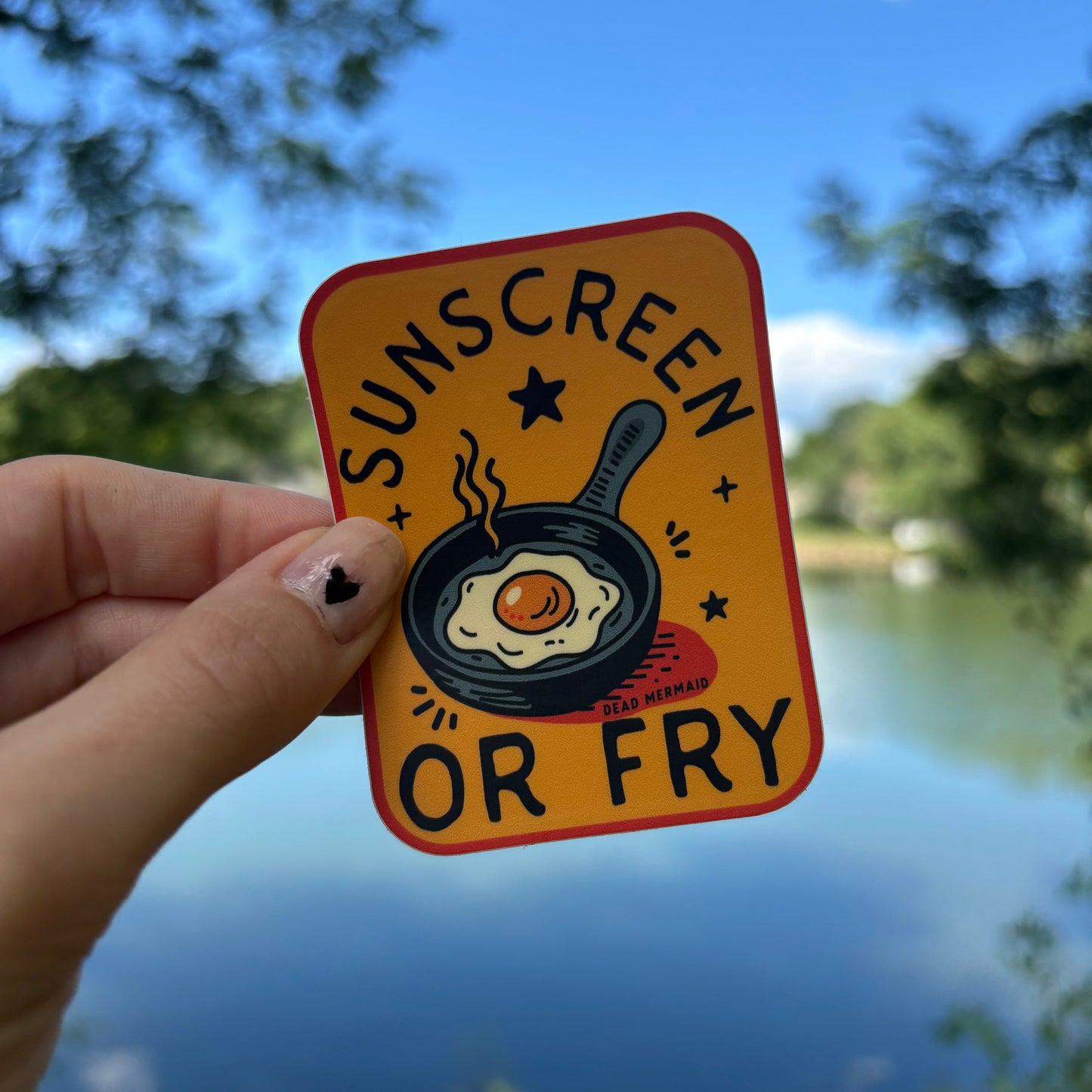 Hand holding a square sticker with a frying pan and egg illustration against a blue sky background.