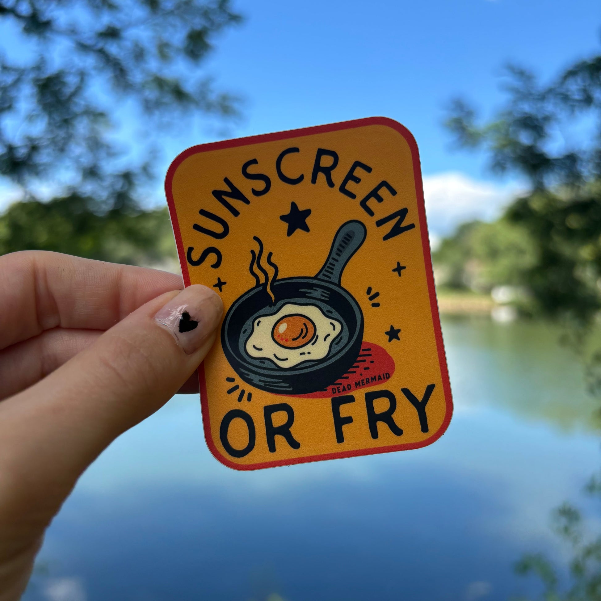 Hand holding a square sticker with a frying pan and egg illustration against a blue sky background. 